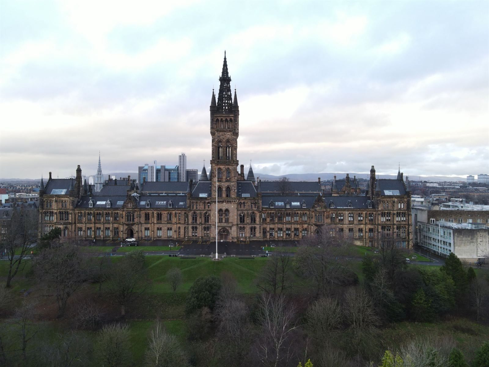The Gothic main building and tall spired tower of the University of Glasgow above a wooded park