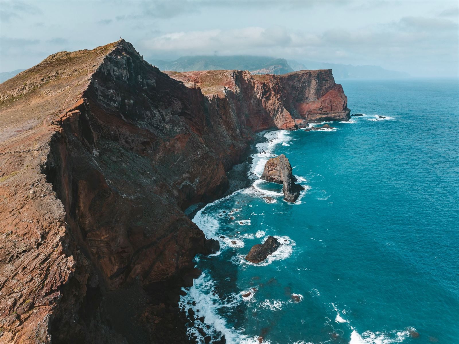 Rugged red and dark volcanic sea cliffs and rock stacks along the Madeira coast, with surf below