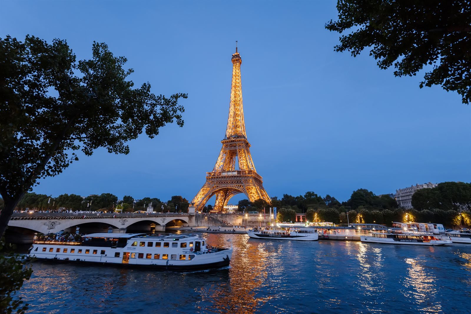 The Eiffel Tower lit up at dusk above the Seine, with tour boats moored along the river