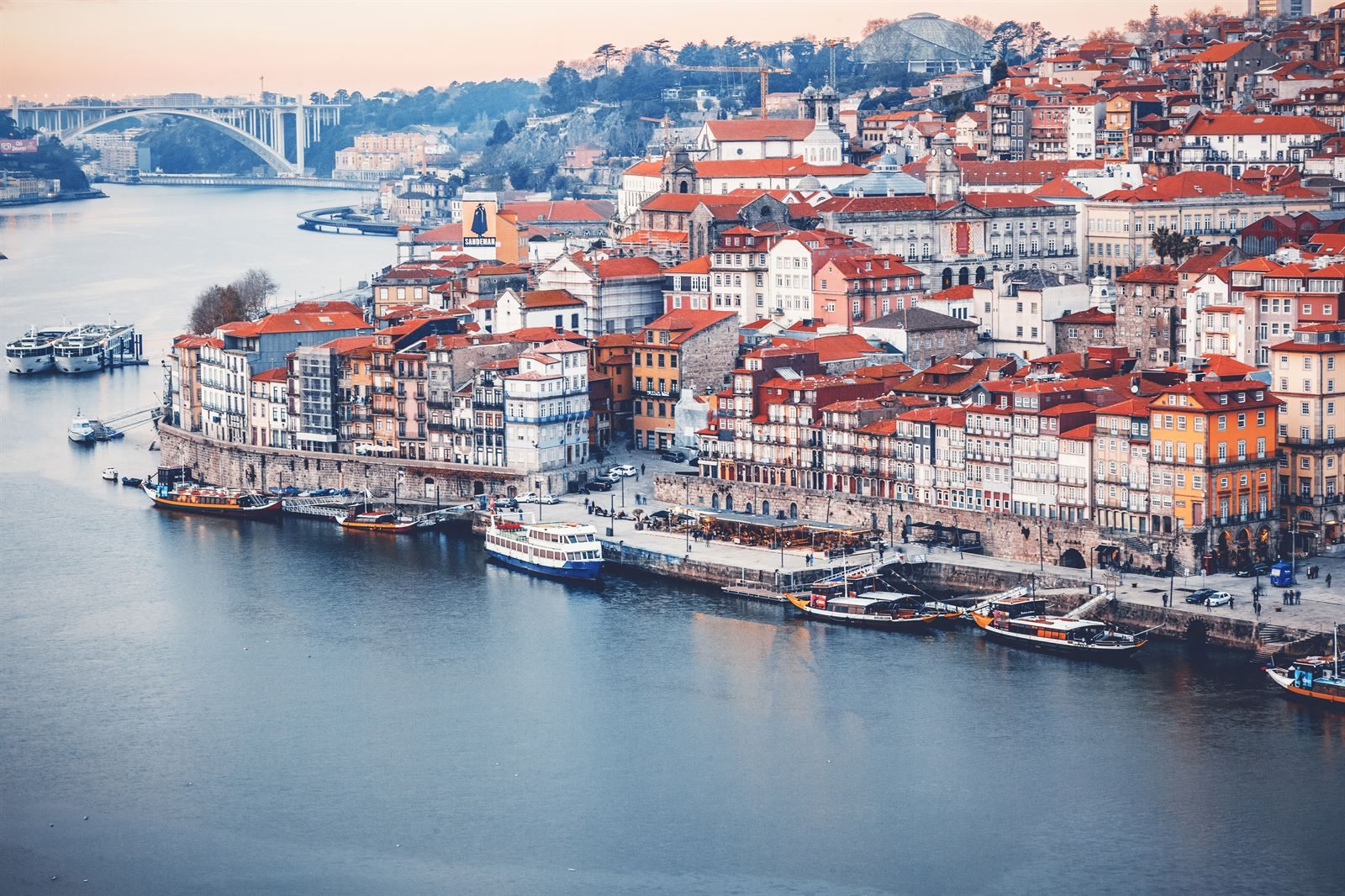 Colorful houses of Porto&rsquo;s Ribeira district on the Douro River, with traditional boats moored below and an arched bridge upstream